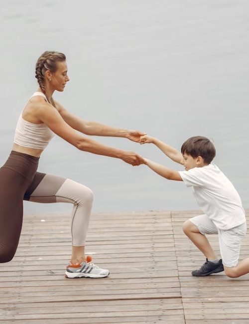 A woman and a boy exercise together on a wooden deck by the lake, promoting healthy lifestyle.
